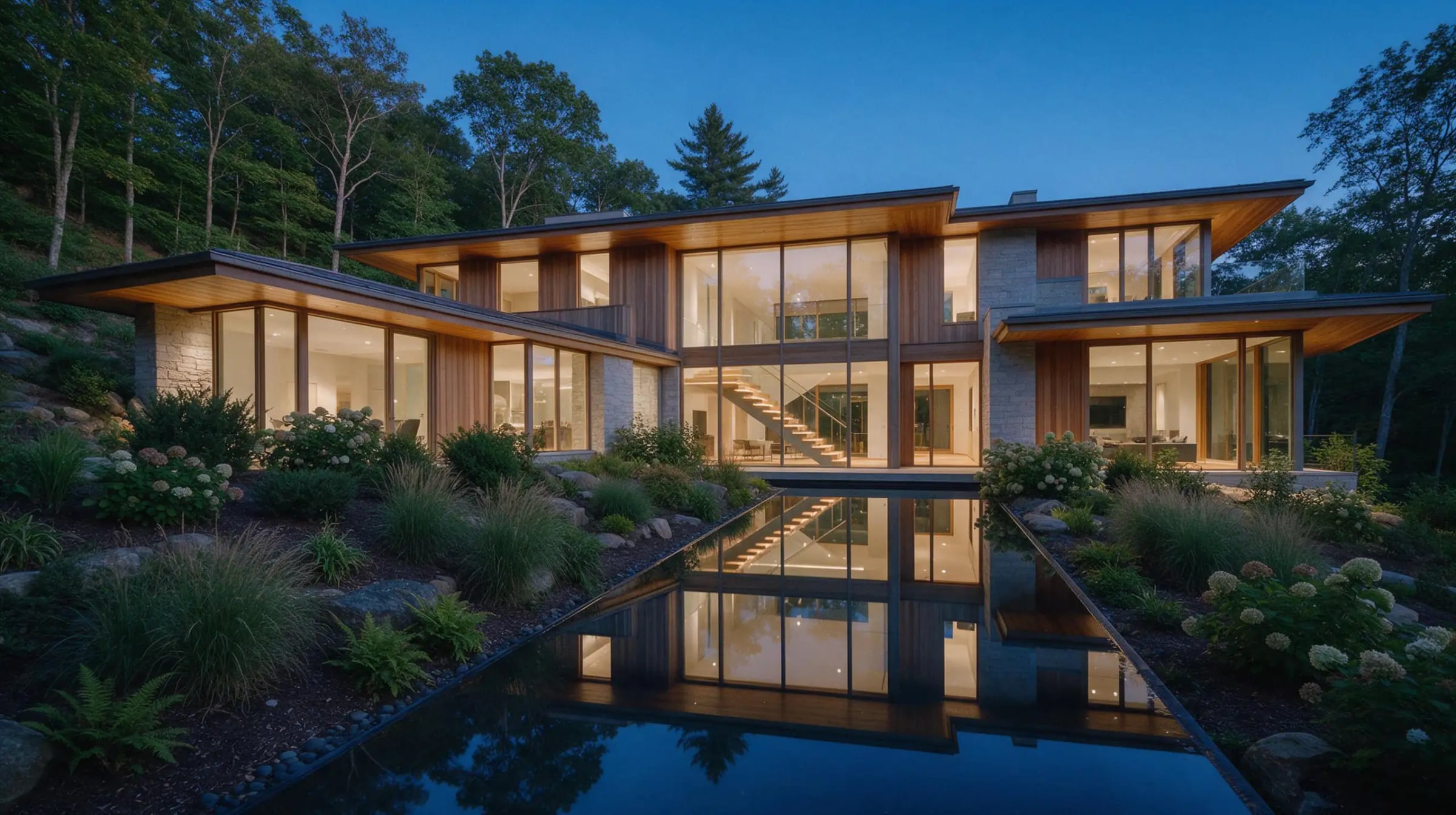 A large modern home of glass, cedar and stone glowing at dusk behind a long reflecting pool and planted terraced garden.