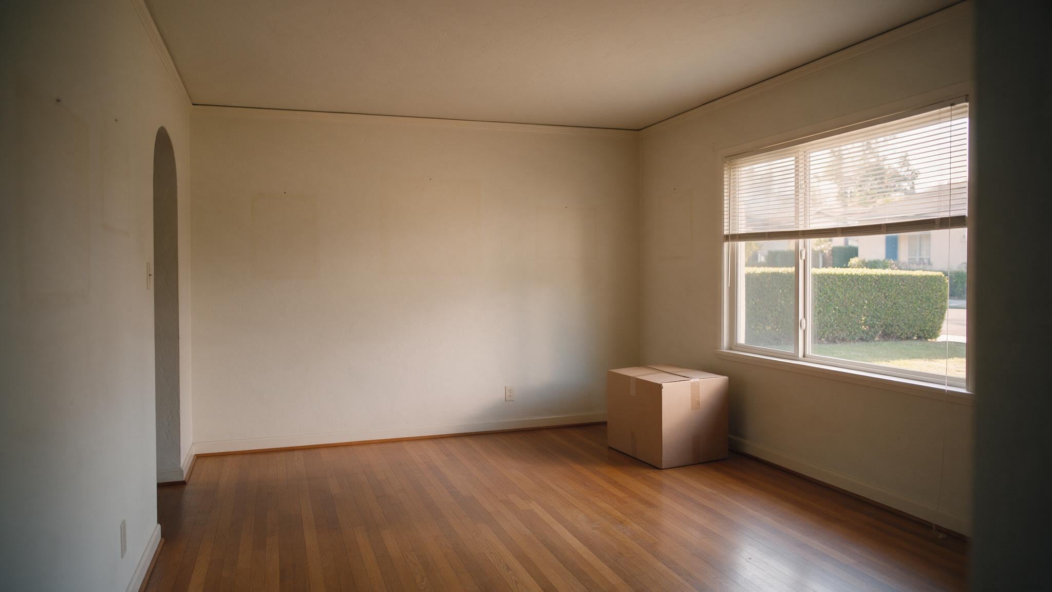Empty room with wooden flooring, beige walls, and bright daylight from large windows overlooking a hedge and street.